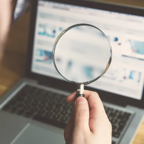 A hand holds a magnifying glass in front of a laptop screen, symbolizing search engine optimization.