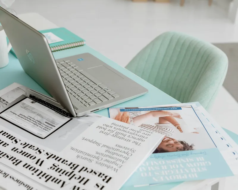 A laptop, magazines, and notebooks for digital marketing are arranged on a mint-colored desk by a green chair.