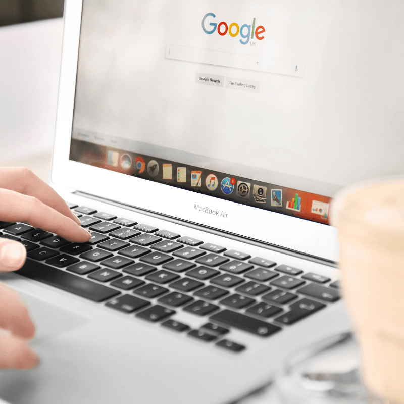 website (3) Person typing on a MacBook Air laptop with the Google homepage open on the screen and a cup in the foreground.
