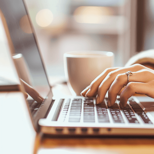 3 Hands typing on a laptop keyboard with a coffee cup in the background on a wooden table.